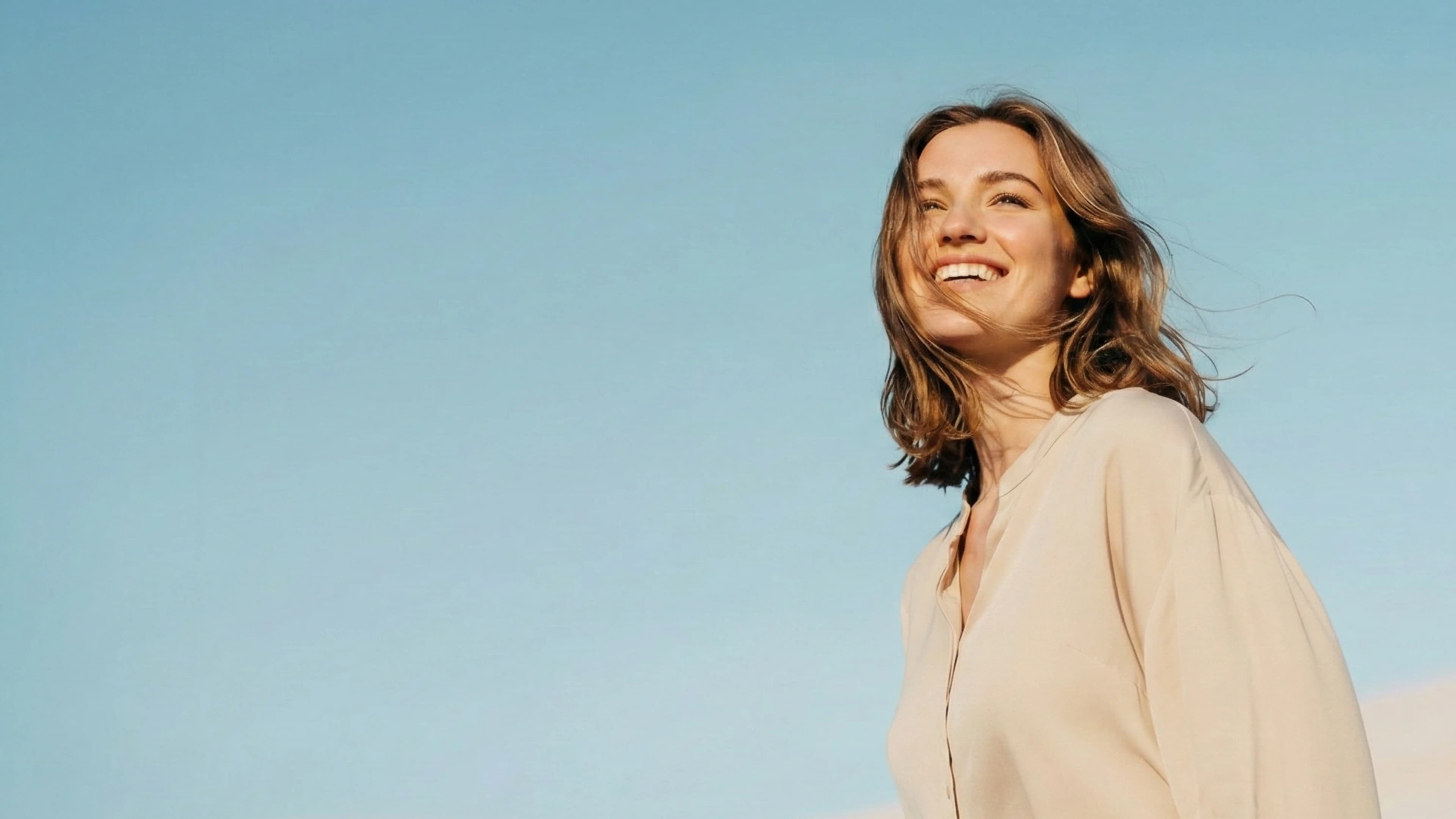 Smiling woman in beige blouse against clear blue sky, looking upward in bright natural light.