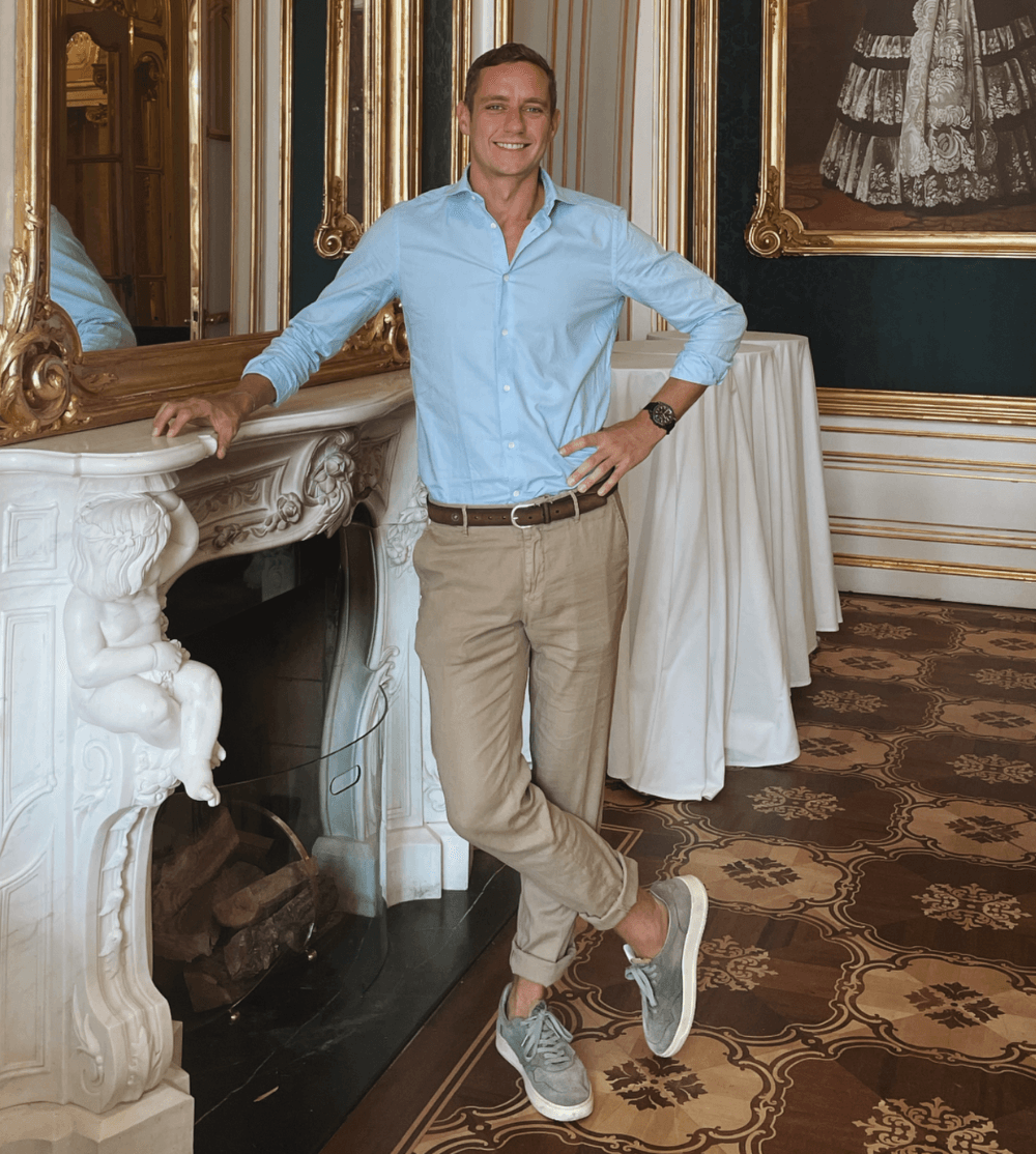 Man in a light blue shirt standing in an ornate interior room with framed art and patterned carpet.