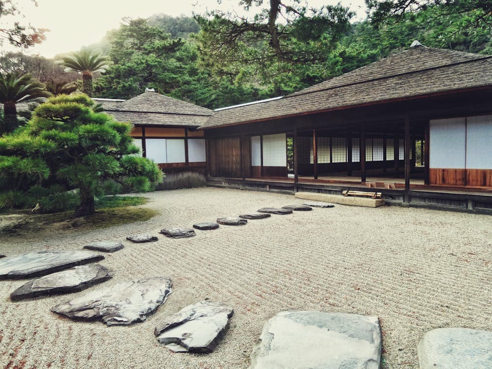 Traditional Japanese garden courtyard with stepping stones, raked gravel, and a wooden building in the background.