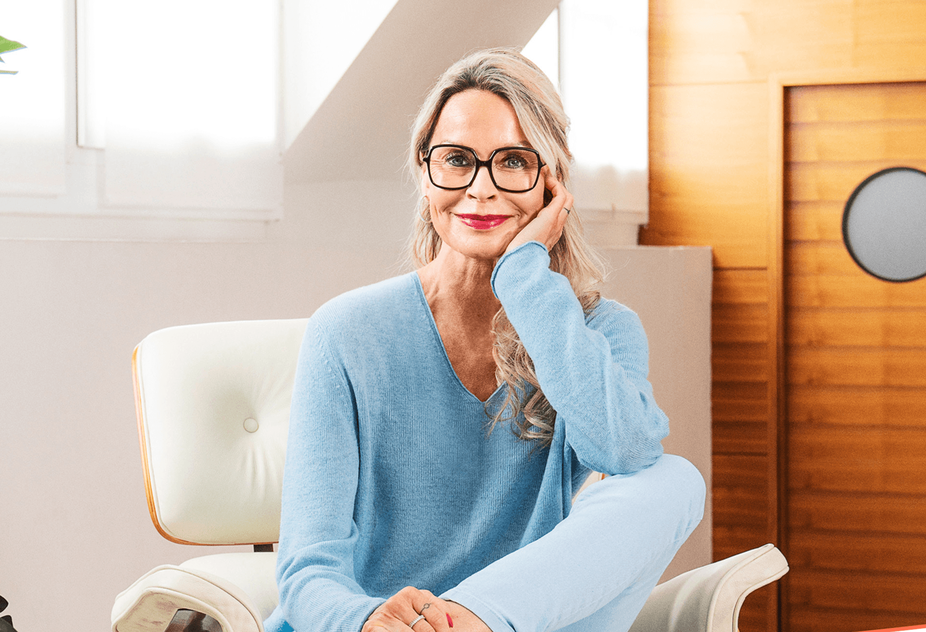 Older woman with glasses in a blue sweater, seated in a chair indoors, smiling with a relaxed pose.