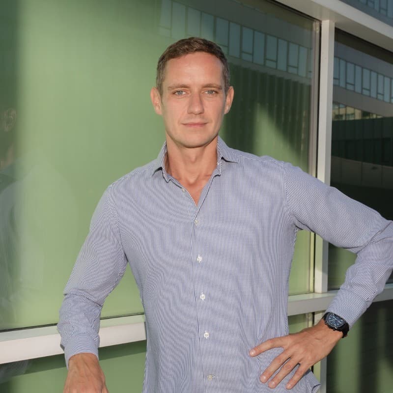 Man in a light shirt leaning on a railing outdoors, with a building facade in the background.
