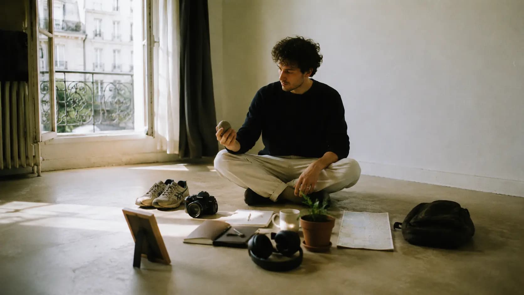 A man with curly hair sitting cross-legged on a bare concrete floor in a minimalist, sunlit room. He is wearing a dark, long-sleeved shirt and light-colored trousers. He looks thoughtfully down at a small, smooth stone he is holding in his right hand.